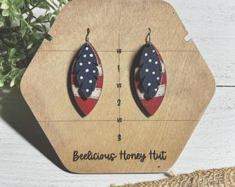 May include: A pair of wooden earrings shaped like a feather with a red, white, and blue American flag design. The earrings are displayed on a wooden board with a ruler and the text "Beelicious Honey Hut".