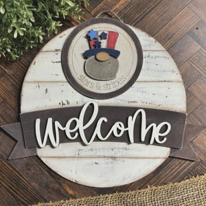 May include: A round wooden sign with a whitewashed finish and a brown banner that says "Welcome". The sign features a red, white, and blue gnome wearing a top hat with stars and stripes. The circle around the gnome says "stars & stripes".