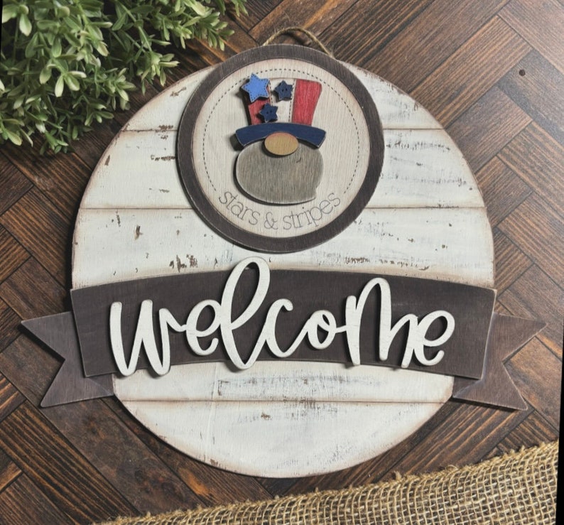 May include: A round wooden sign with a whitewashed finish and a brown banner that says "Welcome". The sign features a red, white, and blue gnome wearing a top hat with stars and stripes. The circle around the gnome says "stars & stripes".