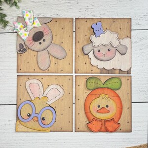 Peut inclure: Quatre plaques carrées en bois avec des motifs sur le thème de Pâques. Les motifs comprennent un lapin avec un nœud, un mouton avec un lapin bleu sur la tête, un poussin portant des lunettes et un canard dans un costume de carotte. Chaque motif est peint dans des couleurs pastel douces.