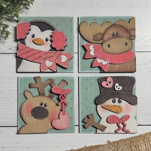 Peut inclure: Quatre décorations de Noël en bois avec un fond vert. Les décorations représentent un pingouin, un élan, un renne et un bonhomme de neige. Chaque décoration a un cœur rouge et un nœud rouge.