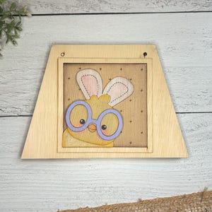 Peut inclure: Une pancarte en bois encadrée avec un poussin jaune portant des lunettes bleues et des oreilles de lapin roses. Le poussin est sur un fond brun avec des étoiles blanches.