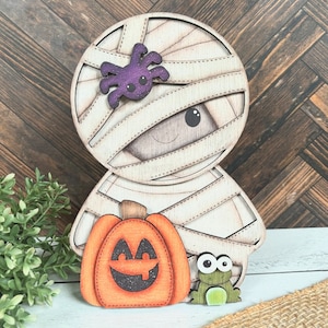 May include: A wooden cutout of a mummy holding a jack-o'-lantern and a frog. The mummy is wrapped in white bandages and has a purple spider on its head. The jack-o'-lantern has a smiling face and the frog has big eyes.