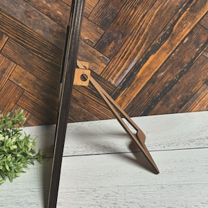 May include: A black wooden frame with a brown easel stand. The frame is standing on a white wooden surface.