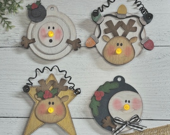 May include: Four wooden Christmas ornaments with light-up faces. The ornaments feature a snowman, a reindeer, a reindeer on a star, and a snowman with a black and white bow.