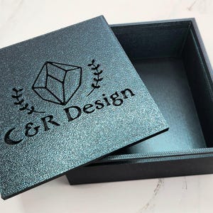 May include: An open, square box with a textured, teal lid. The lid features a geometric design, laurel leaves, and the text "C&R Design" in black. The box interior is a darker teal color.