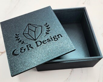 May include: An open, square box with a textured, teal lid. The lid features a geometric design, laurel leaves, and the text "C&R Design" in black. The box interior is a darker teal color.