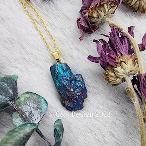 May include: A gold chain necklace with a blue and iridescent crystal pendant. The pendant is a raw, natural crystal with a rough, irregular shape. The necklace is photographed on a white background with dried flowers and green leaves.