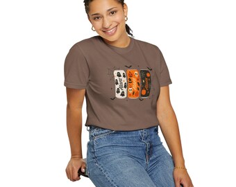May include: A brown t-shirt featuring a Halloween-themed design. The graphic includes three stylized beverage cans with spooky illustrations of ghosts, jack-o'-lanterns, and bats. The word "Alani" is printed on each can.