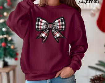 May include: Maroon sweatshirt featuring a large bow design. The bow has a plaid pattern in red, black, and white, with a sparkling center. The word "Maroon" is printed in a decorative font at the top right.