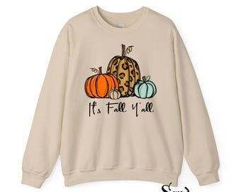 May include: A sand-colored crewneck sweatshirt featuring a graphic of four pumpkins in orange, leopard print, white, and light blue. Below the pumpkins, the text reads "It's Fall Y'all" in a black cursive script. The word "Sand" appears in the bottom right.