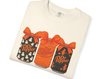 May include: Cream-colored t-shirt featuring three stylized Dr. Pepper cans with orange bows. The cans have Halloween-themed designs: ghosts, jack-o'-lanterns, and witches. The Dr. Pepper logo is visible on each can.