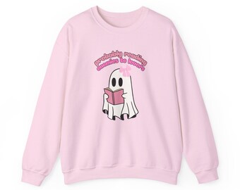 May include: A light pink sweatshirt featuring a cartoon ghost reading a pink book. The ghost has a pink bow and the text "probably reading enemies to lovers" in pink above it. The sweatshirt is a crew neck style.