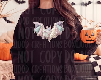May include: Black sweatshirt featuring a colorful bat applique with floral and pastel wing details. The bat design is centered on the chest. A pumpkin-shaped mug is held in the hand. Halloween-themed apparel.