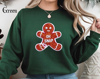 May include: A dark green sweatshirt featuring a glittery red gingerbread man with the words "OH SNAP" in white. The word "Green" is printed in a cursive font in the upper left corner. The sweatshirt is a festive holiday item.