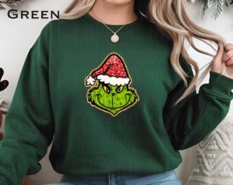 May include: A dark green sweatshirt featuring a graphic of a green character wearing a red Santa hat with glitter accents. The word "GREEN" is printed in white on the upper left. A festive holiday-themed apparel item.