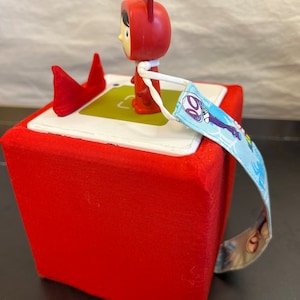 May include: Red fabric cube with a red and white character figurine on top. The cube has a blue and white strap attached to it.