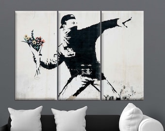 May include: A triptych canvas print featuring a black and white stencil artwork of a figure throwing a bouquet of flowers. The figure is wearing a cap and face covering. The artwork is displayed above a black sofa with white pillows.