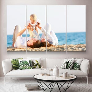 May include: A four-panel canvas print showing a family on a beach. The image features a couple lying on the sand, with a child being held up between them. The background shows the sea and sky. The print is displayed above a white sofa.