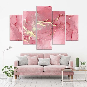 May include: A five-piece abstract art print set featuring pink and gold marble-like patterns. The prints are arranged in a horizontal row on a white wall above a pink sofa.