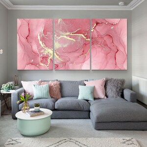 May include: Three-piece abstract art print set featuring pink and gold marble-like patterns. The prints are displayed on a wall above a gray sectional sofa with pillows.