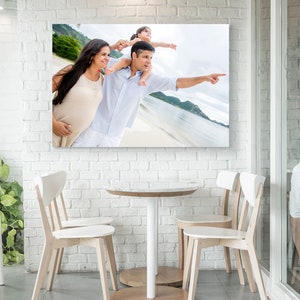 May include: A framed canvas print showing a family on a beach, with a pregnant woman, a man pointing, and a child on his shoulders. The print hangs on a white brick wall above a small round table and chairs.