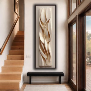 Può includere: Un'opera d'arte murale astratta incorniciata, con un design tridimensionale fluido color crema. L'opera è montata su uno sfondo neutro all'interno di una cornice marrone scuro, creando un contrasto visivo sorprendente. Il pezzo è esposto sopra una panca nera.