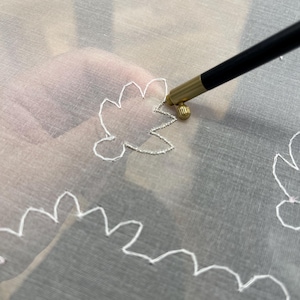 Op de afbeelding: Een close-up van een hand die een zwart en gouden borduurwerktool gebruikt om een wit geborduurd ontwerp op een doorschijnende witte stof te traceren.