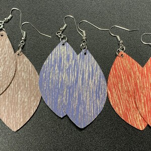 May include: Three pairs of dangle earrings with silver hooks. The earrings are made of leather with a wood grain pattern and a metallic sheen. The colors are a light brown, a light blue, and a reddish brown.