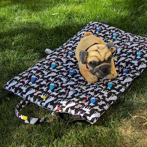 May include: A tan pug plush toy rests on a black and white dog-patterned mat with a black handle. The mat is rectangular and placed on green grass. The dog plush has a red collar.