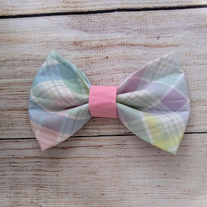 May include: A pastel plaid bow tie with a pink center band. The bow tie features a pattern of light blue, green, pink, and purple squares. The bow tie is made of fabric and is likely for a pet or child.
