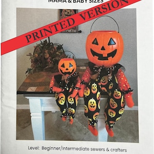 May include: A printed pattern for two pumpkin pail shelf-sitter dolls in mama and baby sizes. The dolls feature orange plastic pumpkin heads with black facial features, black ruffled collars, and fabric bodies with a black pumpkin print pattern.