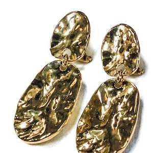 May include: A pair of gold-toned drop earrings featuring two connected, hammered-texture oval discs. Each earring consists of a smaller oval top piece linked to a larger, elongated oval bottom piece, both with a reflective, uneven surface.