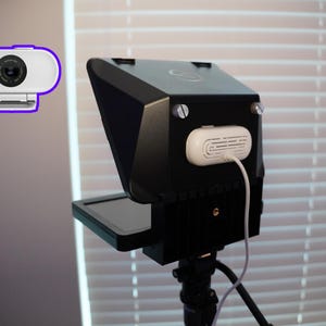 May include: A black teleprompter with a small screen and a white webcam with a purple outline. The teleprompter has a white device attached with a white cable. The setup is on a tripod.