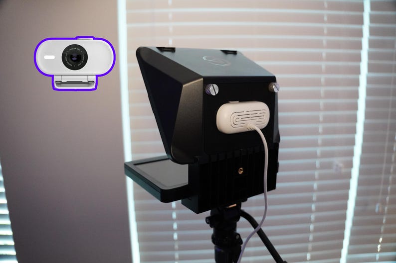 May include: A black teleprompter with a small screen and a white webcam with a purple outline. The teleprompter has a white device attached with a white cable. The setup is on a tripod.