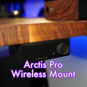 Op de afbeelding: Een zwarte SteelSeries Arctis Pro Wireless headsetbevestiging bevestigd aan een houten bureau. De bevestiging heeft een volumeknop en een zwarte basis.