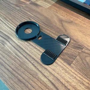 Puede incluir: Un soporte para tel&eacute;fono de color negro impreso en 3D con tiras adhesivas en la parte posterior. El soporte est&aacute; dise&ntilde;ado para sujetar un objeto redondo, como un tel&eacute;fono o un altavoz.
