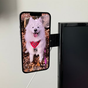 Puede incluir: Un tel&eacute;fono inteligente blanco con una funda negra est&aacute; montado en el lateral de un televisor negro. La pantalla del tel&eacute;fono muestra una foto de un perro blanco con un pa&ntilde;uelo rojo y negro a cuadros. La hora que se muestra en la pantalla del tel&eacute;fono es 9:58.