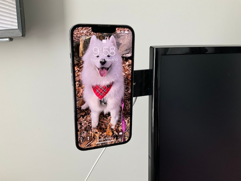 Puede incluir: Un tel&eacute;fono inteligente blanco con una funda negra est&aacute; montado en el lateral de un televisor negro. La pantalla del tel&eacute;fono muestra una foto de un perro blanco con un pa&ntilde;uelo rojo y negro a cuadros. La hora que se muestra en la pantalla del tel&eacute;fono es 9:58.