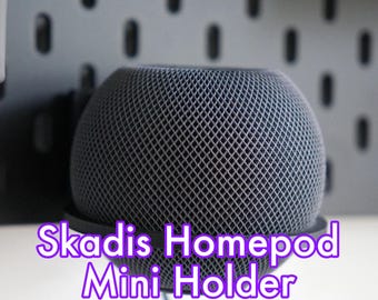 Puede incluir: Un altavoz HomePod Mini redondo de color gris está montado en una estantería de pared Skadis negra. El texto "Skadis Homepod Mini Holder" está impreso en morado debajo del altavoz.