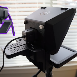 May include: A black webcam and teleprompter setup. The webcam is mounted on a stand, and the teleprompter has a screen. The setup is designed for video recording and streaming.