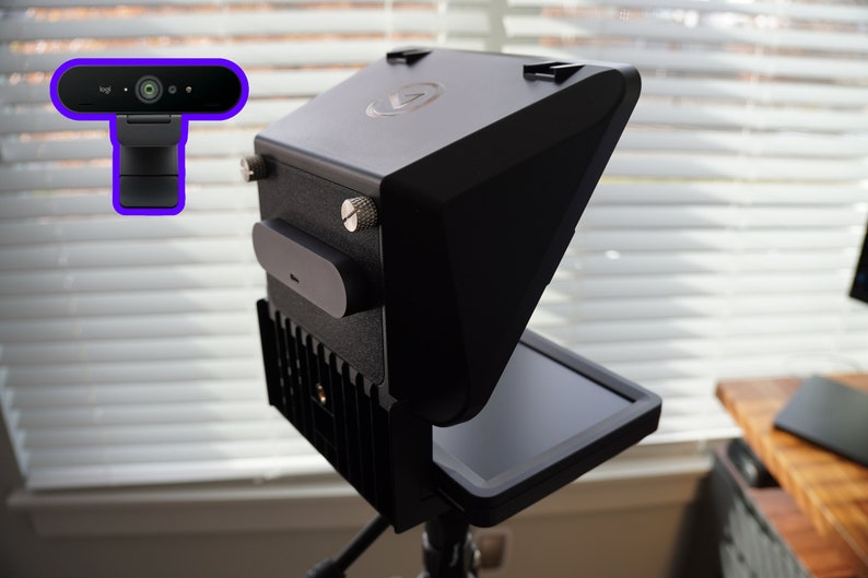 May include: Black video recording equipment with a built-in screen and a separate webcam. The device is mounted on a tripod and has a sleek, modern design. The background features a window with blinds.