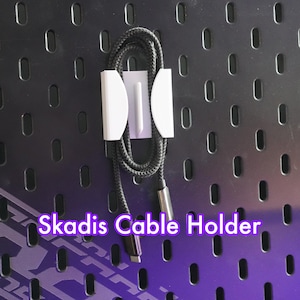 以下が含まれることがあります： 白いプラスチッククリップが付いた黒いケーブルホルダー。ケーブルホルダーは穴のある黒いペグボードに取り付けられています。ケーブルホルダーの下に紫色の文字で「Skadis Cable Holder」と印刷されています。