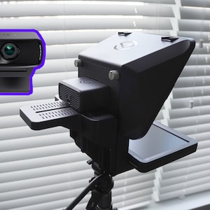 May include: A black webcam and a black teleprompter setup. The webcam is rectangular with a lens and a small light. The teleprompter has a large screen and is mounted on a tripod. The background is a window with white blinds.