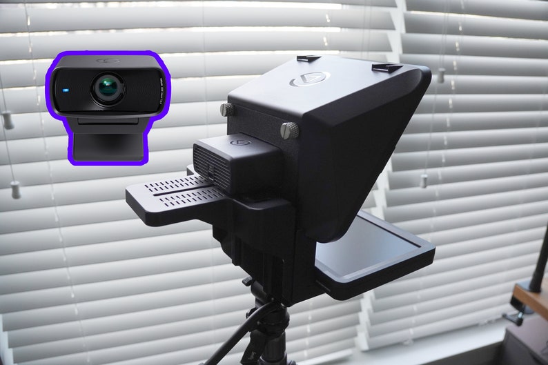 May include: A black webcam and a black teleprompter setup. The webcam is rectangular with a lens and a small light. The teleprompter has a large screen and is mounted on a tripod. The background is a window with white blinds.