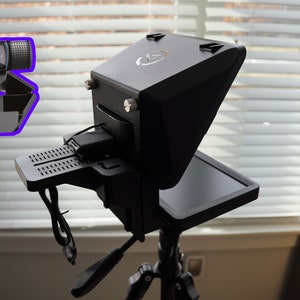 May include: Black teleprompter device with a built-in screen, mounted on a tripod. A webcam is attached to the top. The device has a sleek, modern design, with a matte finish. The background includes a window with blinds.
