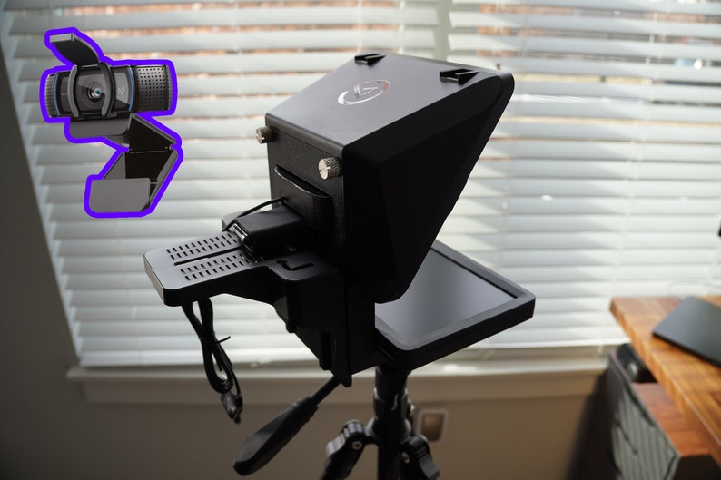 May include: Black teleprompter device with a built-in screen, mounted on a tripod. A webcam is attached to the top. The device has a sleek, modern design, with a matte finish. The background includes a window with blinds.