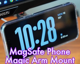 Puede incluir: Una funda transparente para teléfono con un teléfono negro en su interior. La pantalla del teléfono muestra la hora 10:28 y la fecha LUN 16. El texto "MagSafe Phone Magic Arm Mount" se muestra debajo del teléfono.