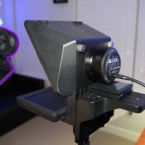 May include: A black webcam with a round lens and a built-in stand is shown in the image. The webcam is mounted on a larger, gray device with a screen and a black base. A black cable is connected to the device.