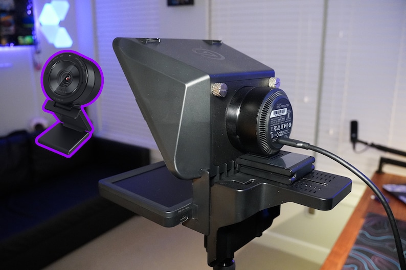 May include: A black webcam with a round lens and a built-in stand is shown in the image. The webcam is mounted on a larger, gray device with a screen and a black base. A black cable is connected to the device.